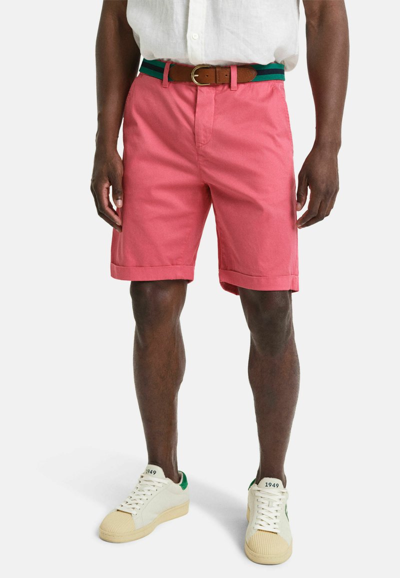 Roze katoenen shorts met een omgeslagen zoom, een bruine leren riem met groene en blauwe strepen, gecombineerd met witte sneakers met groene accenten.
