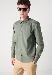 Cămașă verde cu guler clasic, dintr-un material ușor, având nasturi negri. Este combinată cu pantaloni bej și ochelari de soare. Accent de etichetă roșie.