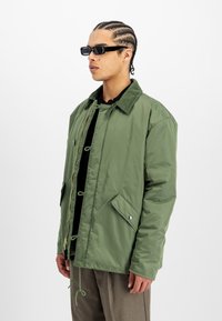 Bărbat purtând ochelari de soare negri, jachetă verde captusită cu guler din bumbac, și pantaloni maro, stând în fața unui fundal alb.