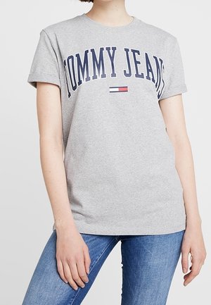 Person wearing a light gray Tommy Jeans T-shirt with navy blue text and logo, paired with blue jeans, standing against a plain background.
