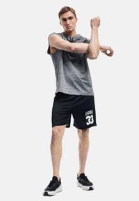 Graues ärmelloses Sportshirt, schwarze Shorts mit dem Aufdruck "LEGENDARY 33" und schwarze Sneakers. Das Modell streckt einen Arm aus und steht vor einem weißen Hintergrund.