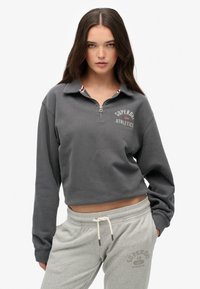 Sweat-shirt gris court avec un col zippé, arborant un imprimé logo, associé à un pantalon de survêtement gris clair avec cordon de serrage et détail imprimé.