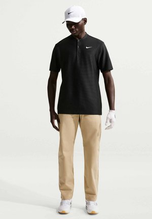 Uomo che indossa una polo nera a maniche corte, pantaloni beige, guanto da golf bianco, berretto bianco e scarpe da ginnastica bianche, in piedi su uno sfondo semplice.