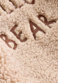 Textil pufos de culoare crem, cu o suprafață texturată, având cuvântul brodat "URSO" în culoare maronie și accente mici albe.