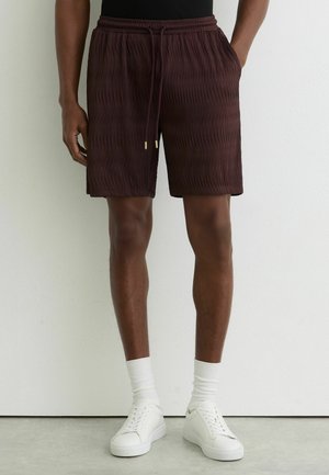 Man draagt donkerbruine, gestructureerde knielange shorts met trekkoord, witte sokken en witte sneakers tegen een effen witte muur.