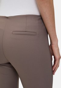 Pantalons taille haute, coupe slim, de couleur taupe avec une texture lisse et une poche arrière unique. Design ajusté avec des coutures visibles.