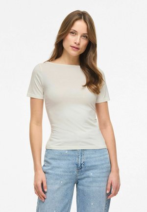 Młoda kobieta z długimi brązowymi włosami, ubrana w dopasowany beżowy krótki rękaw i jasno-niebieskie dżinsy z małymi białymi wzorami kwiatów.