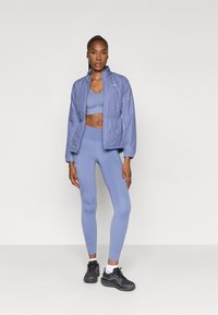 Outfit sportiv de culoare bleu deschis, compus dintr-o jachetă matlasată, un sutien sport ajustat și colanți cu talie înaltă, asortați cu adidași negri.