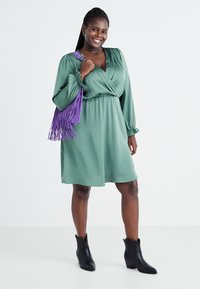 Vihreä, kietaisumallinen mekko, jossa on pitkät hihat ja joustava vyötärö. Yhdistettynä purppuraväriseen fringekassiin ja mustiin nilkkabootseihin. Tasainen pinta.