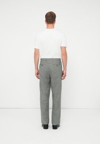 Férfi háttal áll, fehér pólót, szürke kockás nadrágot hátizsák zsebekkel és fekete cipőt visel, egy sima fehér hátt�ér előtt.