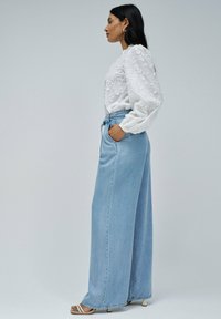 Blouse blanche brodée à manches longues, associée à un jean en denim bleu clair à taille haute et jambes larges. Le modèle se tient de profil.