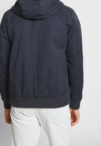 Marinblå, lätt, jacka med huva och ribbad midja och ärmar. Slät yta med synliga sömnadsdetaljer.