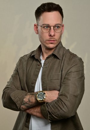 Brown, button-up shirt with rolled sleeves, white T-shirt underneath, and a black and gold watch. Features a tattoo on the forearm.
