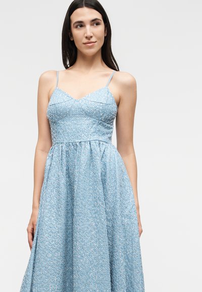 Νέα γυναίκα με μακριά σκούρα μαλλιά, φορώντας ανοιχτού μπλε αμάνικο φόρεμα με λεπτές τιράντες και διακριτικό λευκό μοτίβο, στέκεται σε μονόχρωμο φόντο.