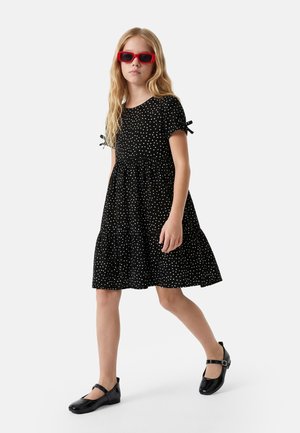 Tyttö kävelee musta pallokuvioinen mekko päällä, mustat Mary Jane -kengät ja punaiset aurinkolasit yksinkertaisen vaalean taustan edessä.