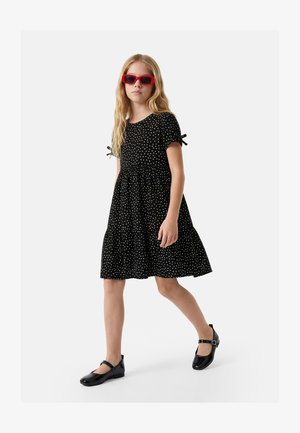 Niña caminando con vestido negro de lunares, zapatos Mary Jane negros y gafas de sol rojas sobre un fondo claro y liso.