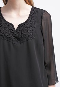 Blusa nera con scollatura a V, caratterizzata da un intricato ricamo nero e bordi a scallop sul petto. Maniche lunghe e trasparenti completano il design.