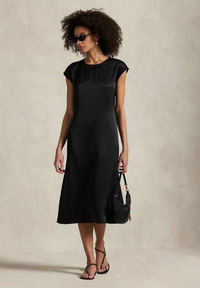 Vestido de cetim preto com mangas tipo capa, comprimento até ao joelho, textura suave. Combinado com sandálias pretas e uma pequena mala, sobre um fundo neutro.