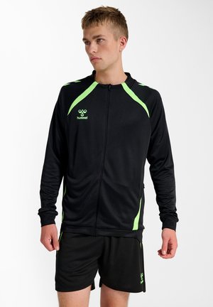 Ung mand iført sort sportjakke og shorts med neongrønne detaljer, stående mod en ensfarvet hvid baggrund.