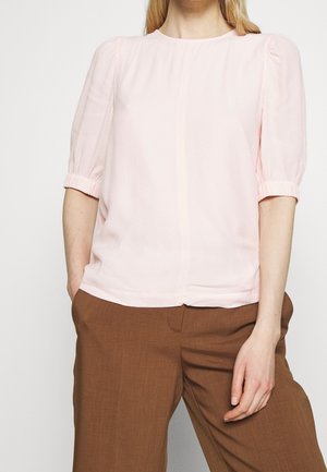 Frau trägt eine hellrosa Bluse mit kurzen, gepufften Ärmeln und braune Hosen, steht mit einer Hand in der Tasche.