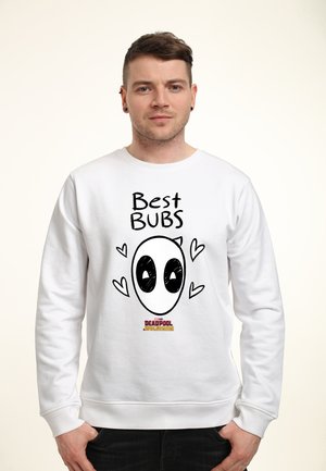 Mężczyzna w białej bluzie z napisem „Best Bubs” i kreskówkową twarzą Deadpoola otoczoną sercami, stojący na jednolitym tle.