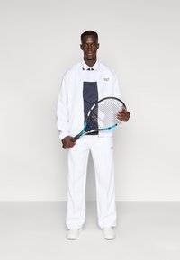 Λευκή φόρμα με ναυτικό μπλε απόχρωση στο πουκάμισο· μοντέλο που κρατά ρακέτα τένις· μινιμαλιστικός σχεδιασμός· απαλή υφή· γκρι λογότυπο στο μπουφάν.