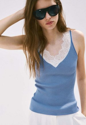 Junge Frau trägt schwarze Sonnenbrille und ein blaues, geripptes Tanktop mit weißer Spitzeneinfassung, steht mit einem Arm hinter dem Kopf erhoben.