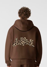 Brun hettegenser med strukturert leopardmønsterdesign på baksiden, med en avslappet passform og stor hette. Enkel, udekoreert front.