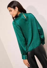 Smaragdrohe satiinbluus, millel on tagaküljel võtmeta detail, pikad varrukad ja nööpidega mansetid. Sujuv sobivus täiendab musti pese.