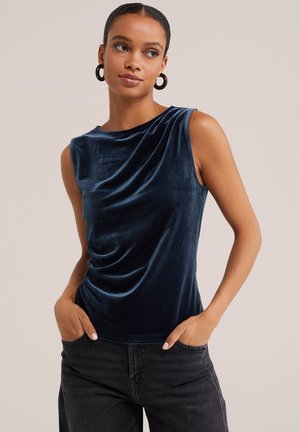 Femme aux cheveux foncés portant des boucles d'oreilles créoles noires, un haut en velours bleu foncé sans manches et un jean noir les mains dans les poches.