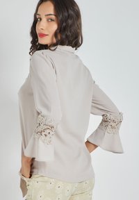 Blusa gris claro con mangas acampanadas, que presenta delicados acentos de encaje en los puños. Textura suave y un diseño simple y elegante.