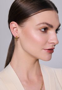 Złota, trójloopowa kolczyk ma polerowane wykończenie. Modelka ma gładkie włosy, nosi jasny sweter, prezentując biżuterię na neutralnym tle.