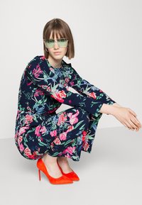 Macacão floral em azul escuro com padrões multicoloridos, complementado por sapatos de salto alto vermelhos e óculos de sol verdes.