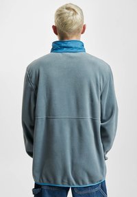 Pullover in fleece grigio con colletto blu, presenta un design senza cuciture e maniche lunghe, mostrando una texture morbida e una vestibilità rilassata.