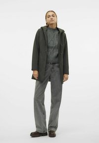 Chaqueta impermeable verde oscura con capucha, superpuesta a un suéter de punto texturizado en gris oscuro, combinada con pantalones vaqueros grises desgastados de pierna ancha y botas marrones.