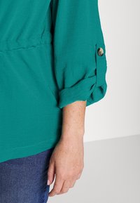 Gr�önblå blus med uppkavlade ärmar och knappdetalj. Mjuk, texturerad tyg med avslappnad passform, matchad med mörkblå denim.