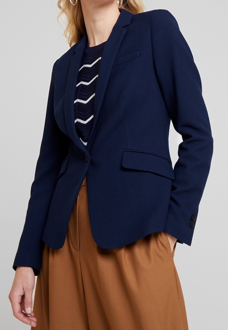 Blazer texturizado azul marino con solapas de muesca, cierre de un solo botón y dos bolsillos frontales, combinado con una blusa a rayas y pantalones marrones.