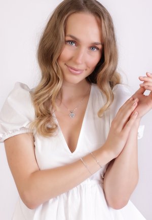 Silver heart pendant necklace and delicate bracelet on a model with wavy hair, wearing a white blouse with puff sleeves against a light background.