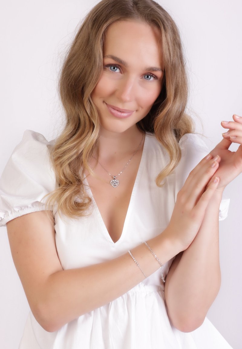 Collier en pendentif cœur en argent et bracelet délicat sur un mannequin aux cheveux ondulés, portant une blouse blanche à manches bouffantes sur fond clair.