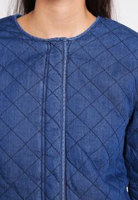 Blå quiltad jacka med rund halsringning, prydd med diamantmönster, framdragkedja och textil med jeanslook.