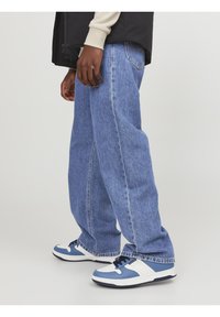 Jean en denim bleu à coupe relaxée avec des coutures visibles, associés à des baskets bleues et blanches avec des lacets noirs et une semelle texturée.