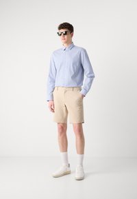 Lys blå og hvit-stripete skjorte med lange ermer, kombinert med beige shorts. Modellen har svarte solbriller og hvite sneakers.