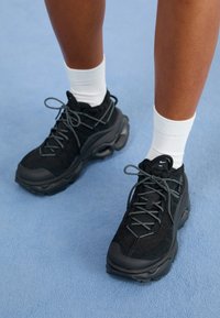 Ben som bär vita strumpor och svarta Nike-sneakers med kraftiga sulor på ett ljusblått texturerat golv.