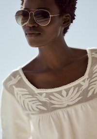 Sunglasses s velkými, reflexními tmavými skly a tenkými rámem z růžového zlata. Bílé tričko s vyšívaným květinovým vzorem a texturovaným výstřihem.