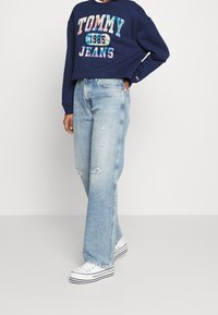 Marinblå croppad sweatshirt med "TOMMY JEANS 1985" grafisk design. Ljusblå vida jeans i utsliten stil. Vita sneakers med svarta ränder.