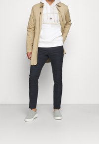 Uomo che indossa un trench beige sopra un hoodie bianco Tommy Hilfiger, pantaloni slim-fit scuri e scarpe slip-on grigio chiaro, in piedi contro uno sfondo bianco.