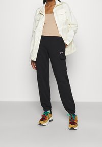 Černé cargo kalhoty se bočními kapsami, doplněné krémovou voděodolnou bundou a béžovým tílkem. Vícebarevné tenisky Nike outfit dokončují.