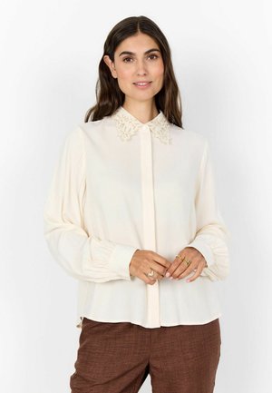 Kobieta o długich brązowych włosach, ubrana w kościelnobiałą bluzkę z koronkowym kołnierzem i bufiastymi rękawami, w połączeniu z brązowymi, teksturowanymi spodniami, stoi z rękami splecionymi.