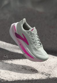 Světle šedá běžecká bota s síťovaným svrškem, růžovými akcenty a texturovanou podrážkou. Má tradiční šněrování a polstrovaný límec.