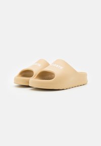 Sandalias sin tacón en color beige, confeccionadas en goma, con un diseño redondeado, punta abierta y logotipo blanco en relieve en la parte superior. Suela texturizada para mayor agarre.